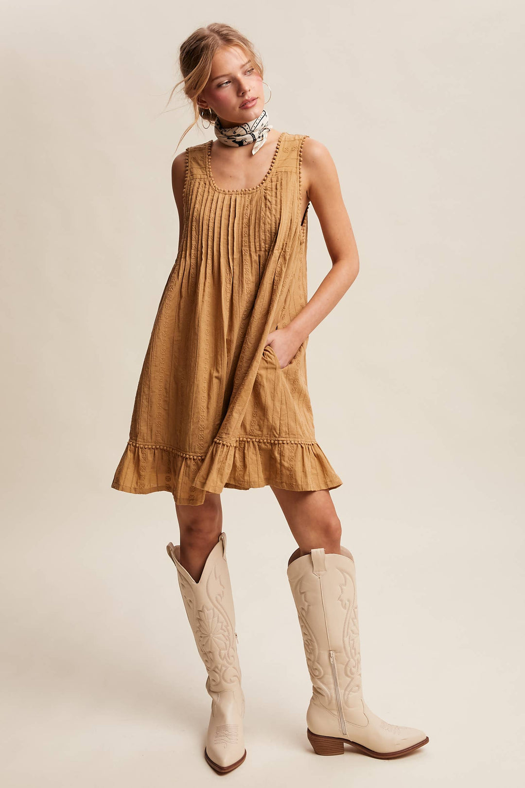Woman wearing a textured camel-colored sleeveless mini dress with pintuck details and ruffle hem.