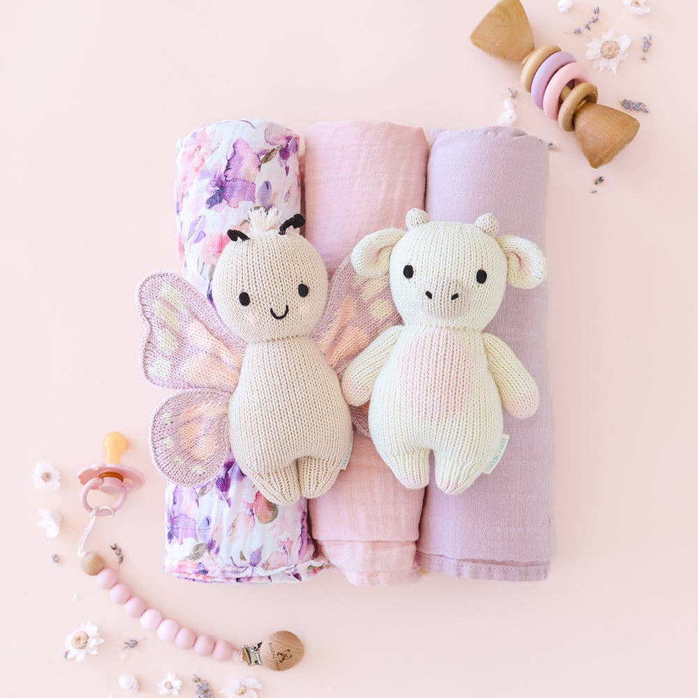 Two knitted toys, a butterfly and a cow , on top of folded baby blankets with a pink background.