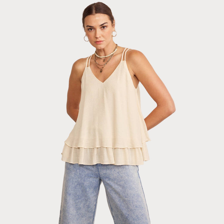 A woman wearing a pink sleeveless blouse with double layered ruffles at the bottom and adjustable shoulder straps. She is also wearing light-colored trousers.