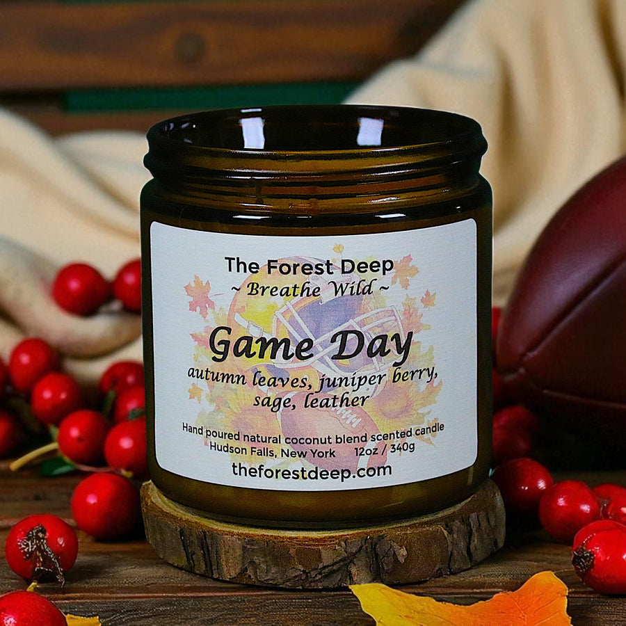 Candle labeled 'Game Day' with autumn leaves, juniper berry, sage, and leather scents on a wooden surface with berries and a football.